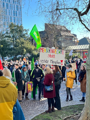 Eine große Menschenmenge versammelt sich im Freien zu einem Protest. Die Menschen halten Schilder und Fahnen hoch, umgeben von Bäumen und städtischen Gebäuden im Hintergrund. (c) Vera Meister